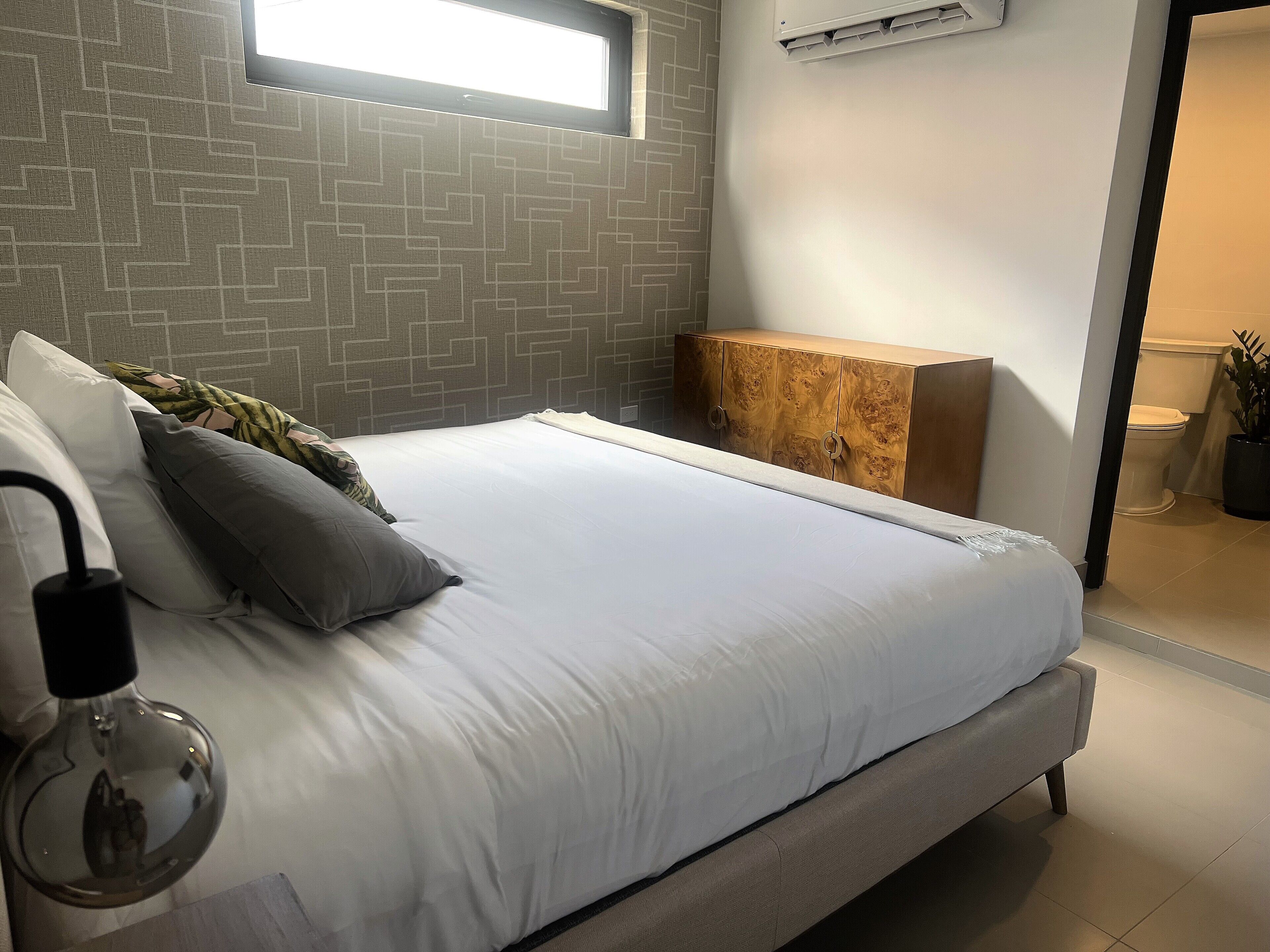 Ein modernes Schlafzimmer mit einem großen Bett, einem Bettrahmen mit Stauraum, einem Fenster und einem durch eine offene Tür einsehbaren Badezimmer.