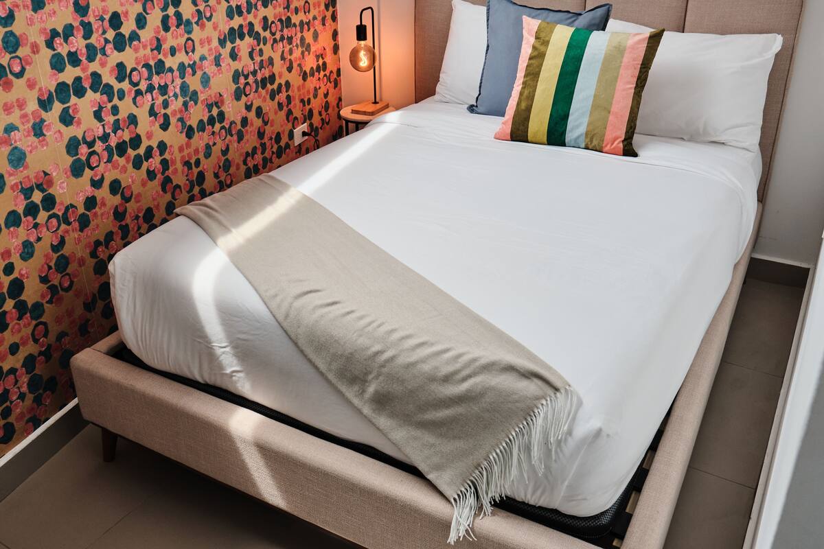 A neatly made bed with a beige throw blanket and multiple pillows, a small bedside table with a lamp, and a wall with a floral pattern.