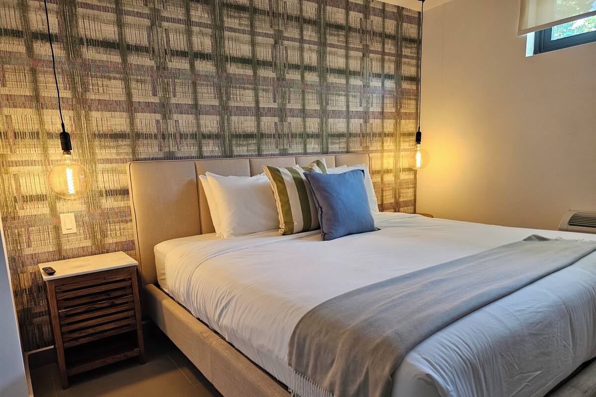 A neatly made bed with a white comforter and pillows, a bedside table with a remote control, and a wall with a textured pattern.