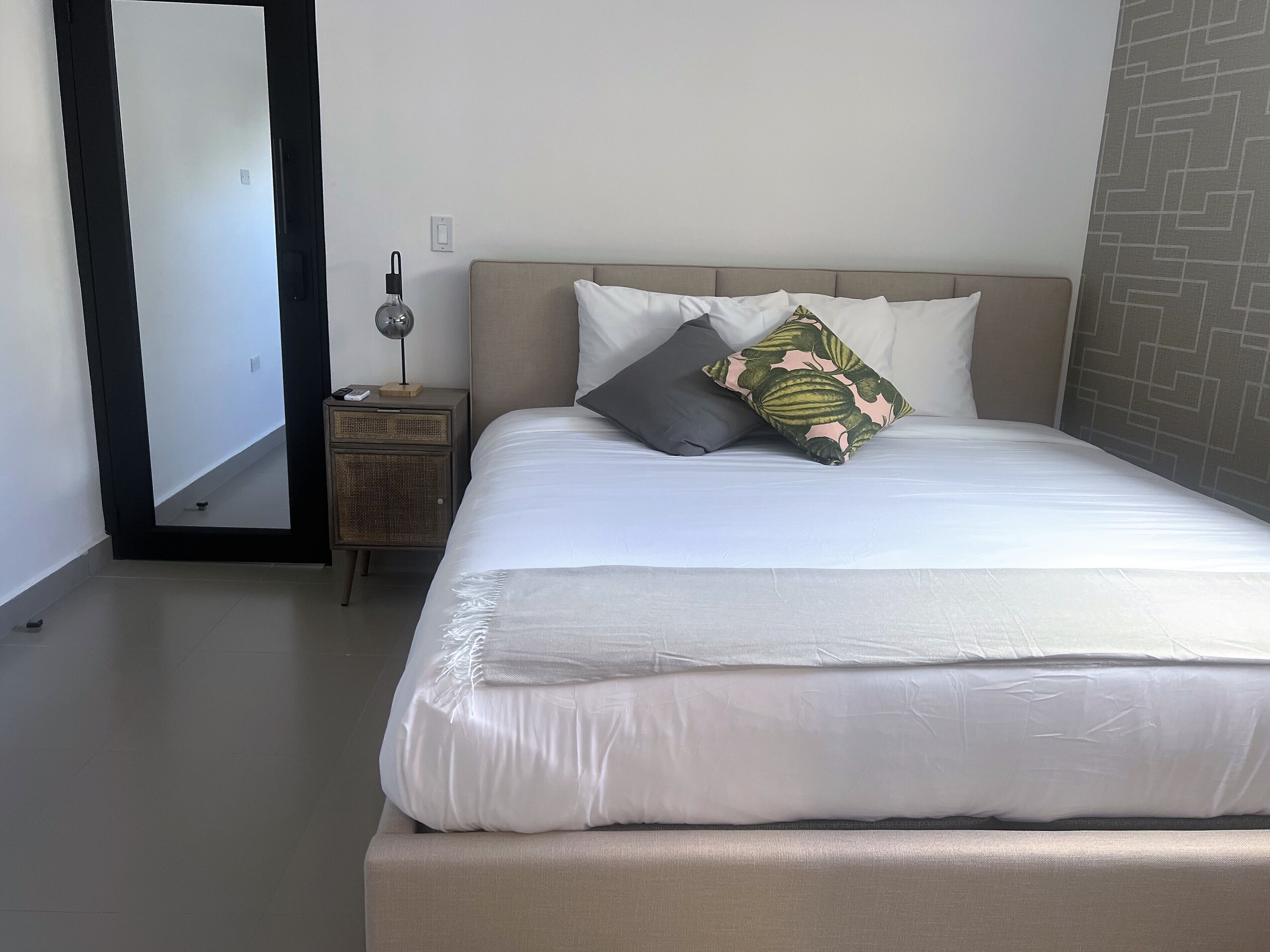 A neatly made bed with white linens and multiple pillows, a bedside table with a lamp, and a wall with a geometric pattern.