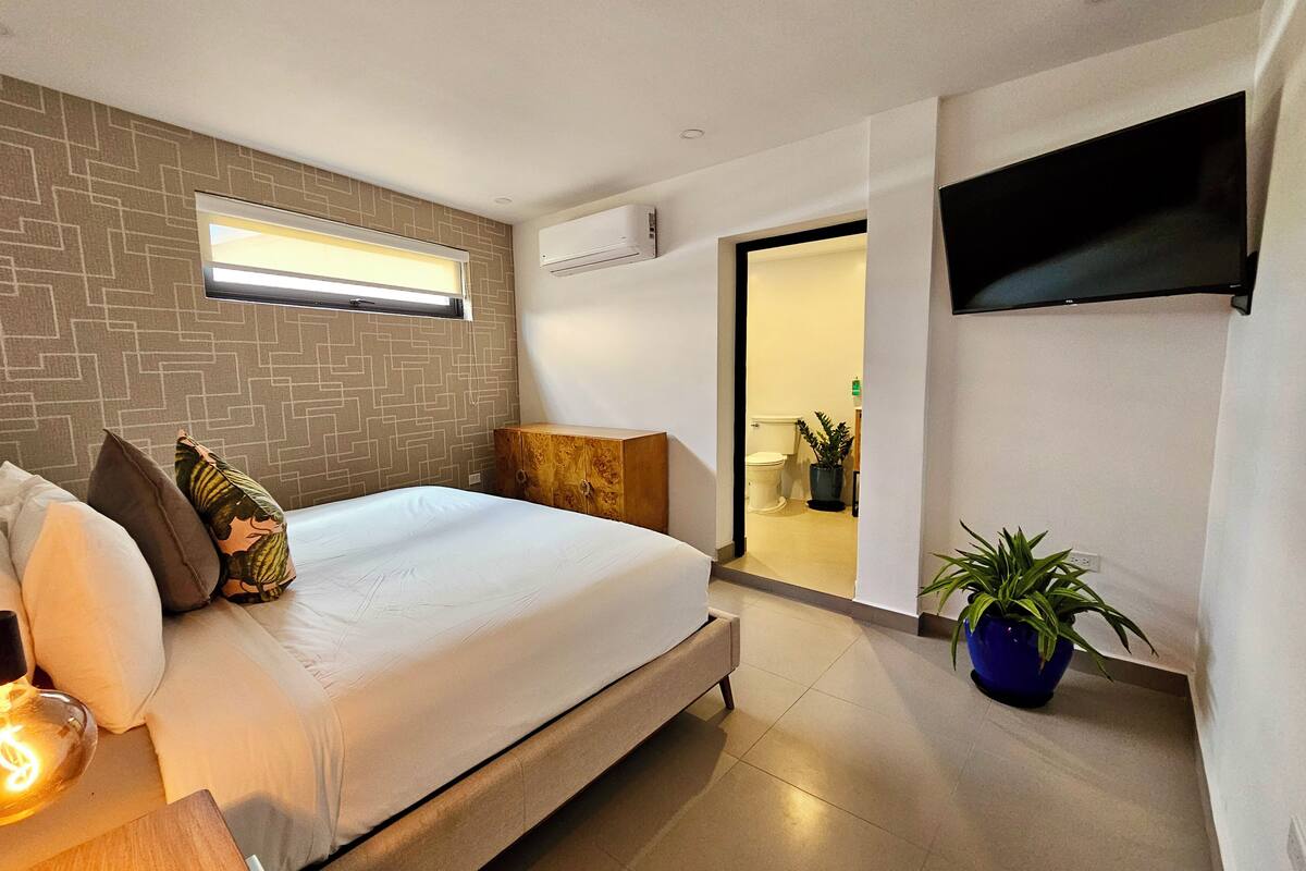 A modern bedroom with a large bed, a wooden nightstand, a wall-mounted TV, and a potted plant.