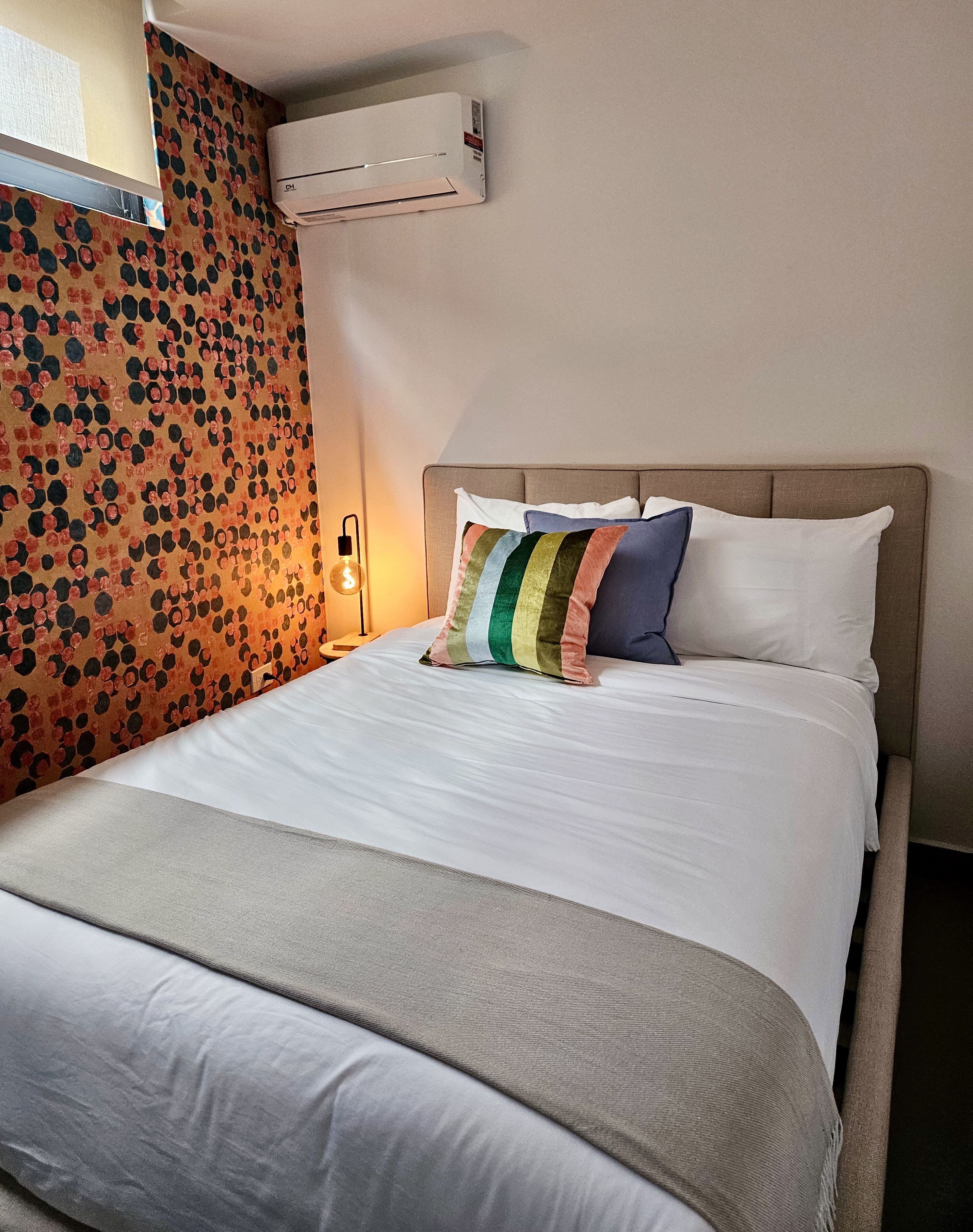 A hotel room with a bed, a wall-mounted air conditioner, and a patterned wall.