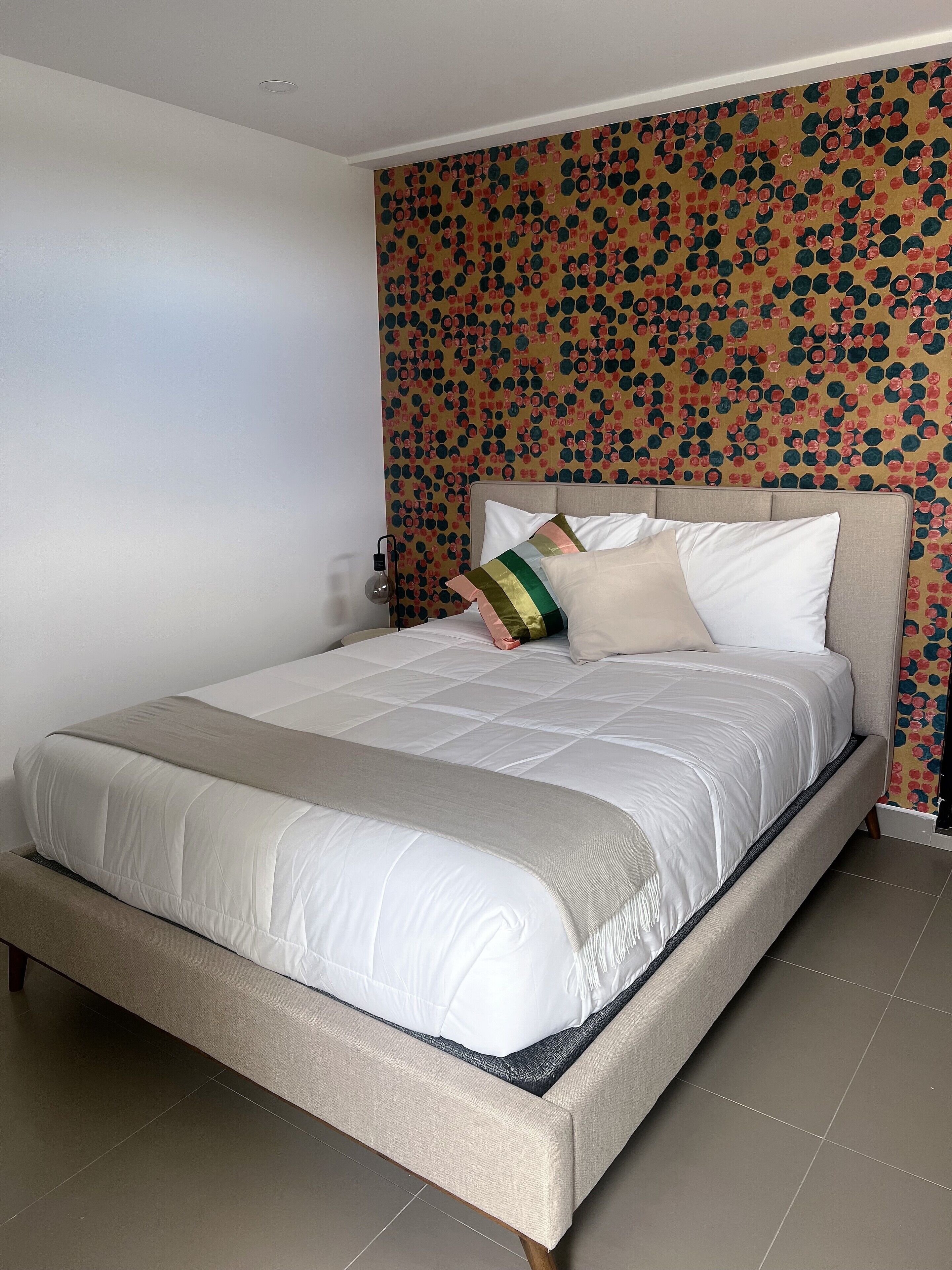 A neatly made bed with a beige headboard and white bedding, a patterned wallpaper, and a small bedside table with a lamp.