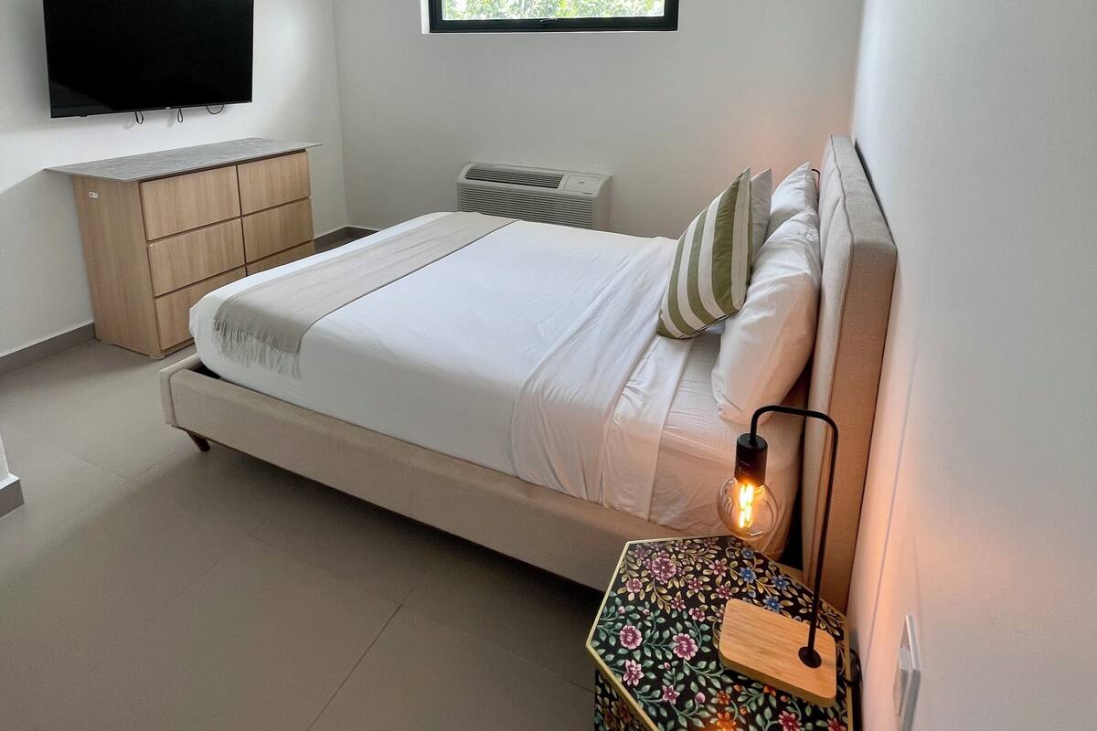 A modern bedroom with a bed, a TV, a window, a bedside table with a lamp, and a decorative box.