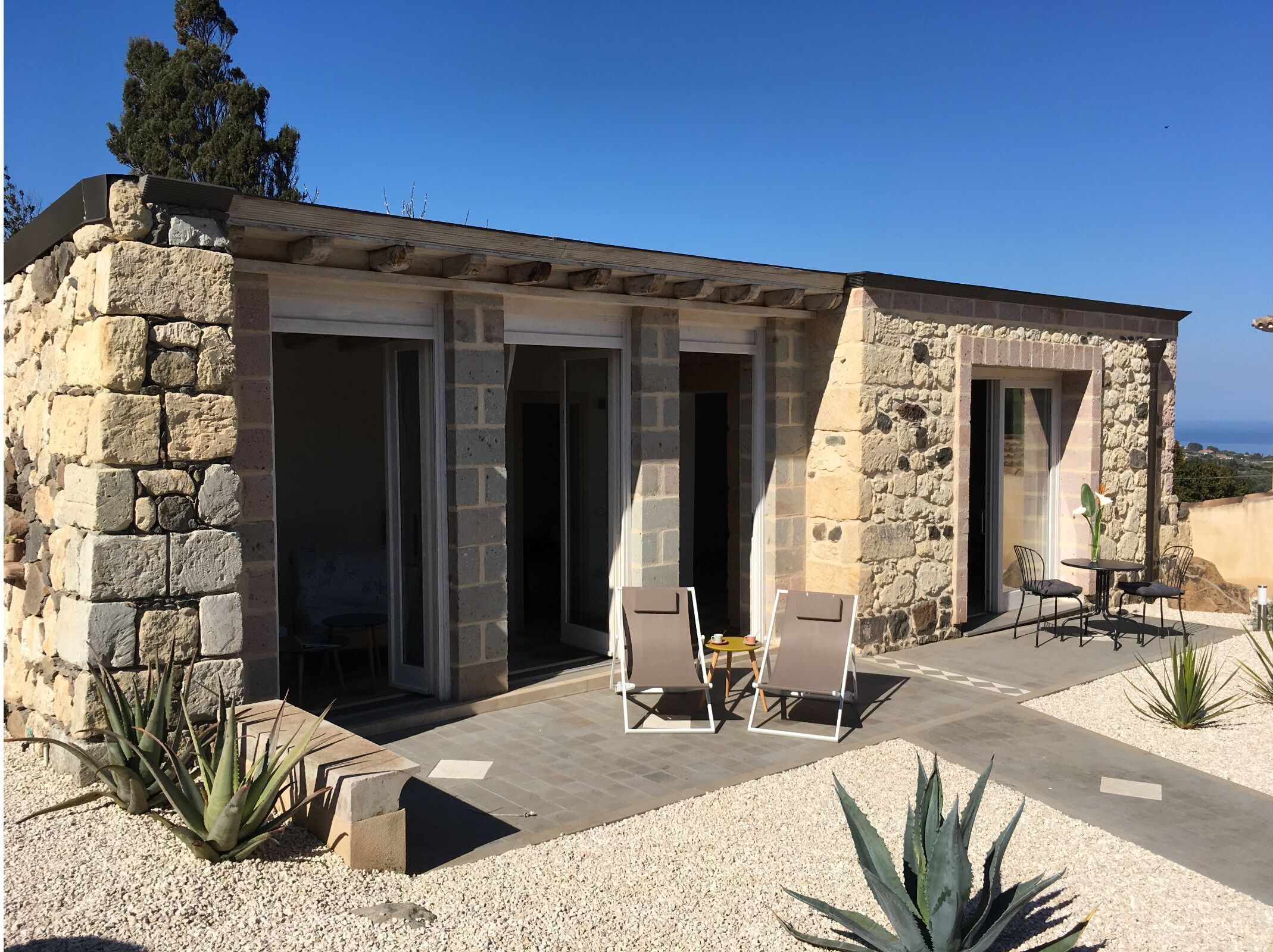A stone building with a patio area featuring two chairs and a table, surrounded by plants and a view of the sea.