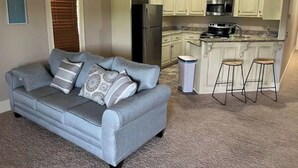 A compact living space with a grey sofa, a kitchen with white cabinets, a stainless steel refrigerator, and a small bar area with stools.