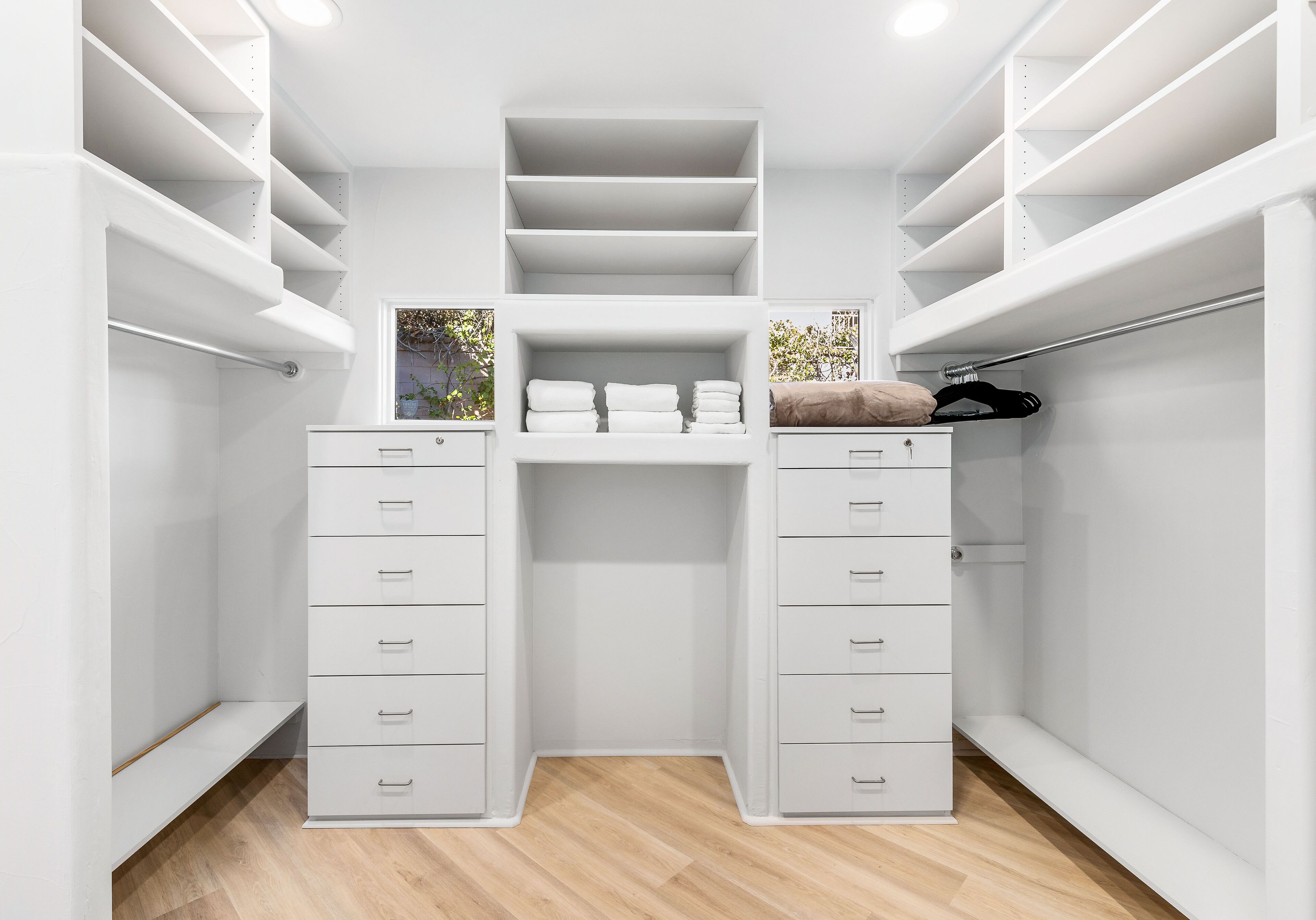 A walk-in closet with multiple shelves and drawers, a wooden floor, and a view of trees outside.