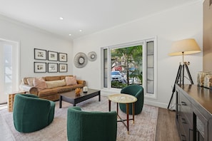 A living room with a sofa, coffee table, and chairs, a lamp, and a view of the street outside.