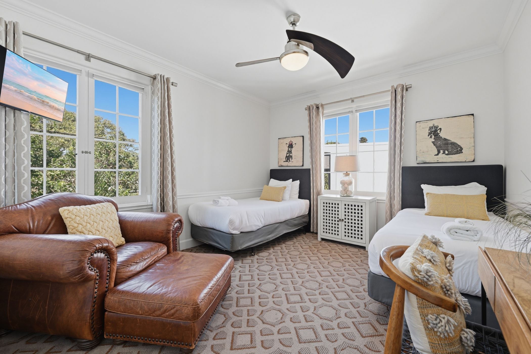 A hotel room with a leather sofa, two beds, a ceiling fan, and artwork on the walls.