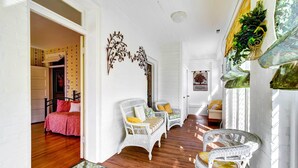 A bright hallway with white walls, wooden flooring, wicker chairs, and a bed with a red bedspread. - Jenny’s Southland Homestead, LLC (Bowling Green)
