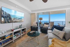 A modern living room with a large TV, comfortable seating, a coffee table, and a view of the ocean.