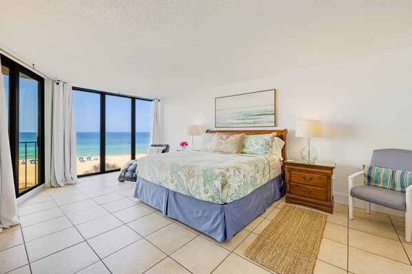 A bedroom with a large bed, a nightstand, a chair, and a view of the beach.