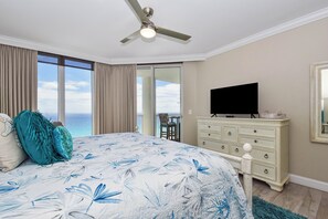 A bedroom with a bed, a ceiling fan, a TV, a dresser, and a view of the ocean.