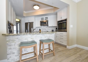 A modern kitchen with white cabinets, a marble backsplash, and stainless steel appliances. There are two wooden stools with green cushions. - Beautifully Updated Silver Beach Towers 3-Bedroom Condo (Destin)