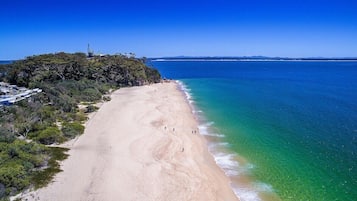 A beach with clear blue water, a sandy shore, and a forested area with a structure on a hill.