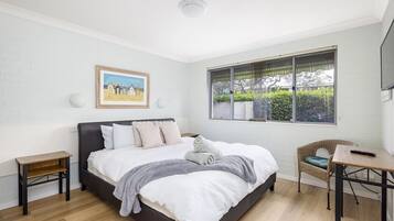 A bedroom with a large bed, a small table, a chair, a window with blinds, and a framed picture on the wall.
