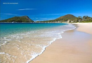 Ein Strand mit klarem türkisblauem Wasser, einem sandigen Ufer und einer hügeligen Landschaft im Hintergrund.