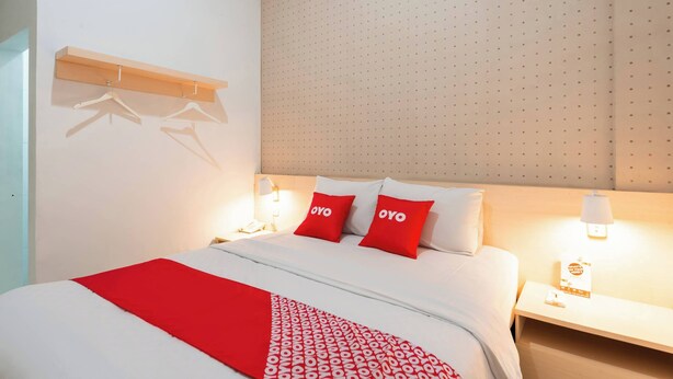 Ein Hotelzimmer mit einem ordentlich bezogenem Bett, zwei Nachttischlampen, einem Wandregal mit Kleiderhaken und einer gemusterten Tagesdecke.