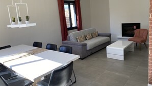 Una sala moderna con un sofá gris, una mesa de centro blanca y una chimenea. Hay una zona de comedor con una mesa blanca y sillas negras.