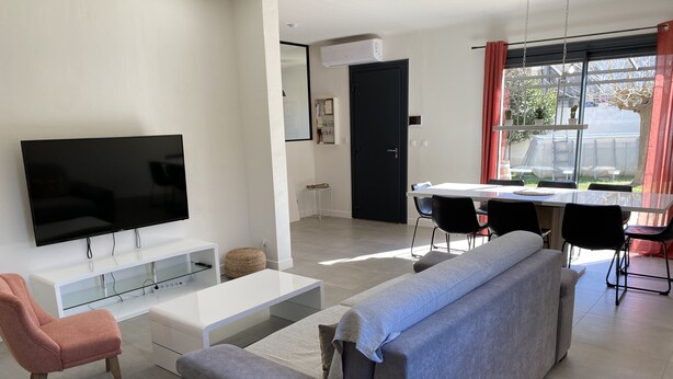 Una sala de estar moderna con un televisor de pantalla plana, un sofá gris, un sillón rosa y una zona de comedor con mesa y sillas.