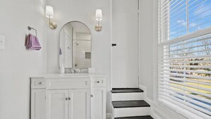 Ein Badezimmer mit weißem Waschtisch, einem Spiegel, zwei Wandleuchten, einem Handtuchhalter und einem Fenster mit Jalousien.