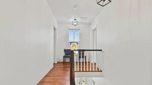 Ein schmaler Flur mit Holzboden führt zu einer Treppe mit schwarzem Geländer. Ein Stuhl steht in der Nähe eines Fensters mit farbigem Glasdekor.