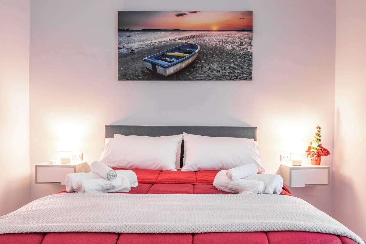 En pænt redt seng med hvidt og rødt sengetøj, et indrammet billede af en båd på en strand og to sengeborde med lamper samt en vase med blomster.