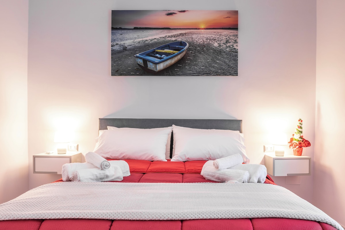 A neatly made bed with white and red bedding, a framed picture of a boat on a beach, and two bedside tables with lamps and a vase of flowers.
