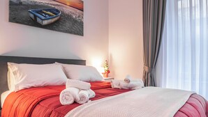 Un letto rifatto con cura, con biancheria rossa, cuscini bianchi e asciugamani arrotolati. Al muro, sopra il letto, un quadro raffigura una barca sull'acqua.