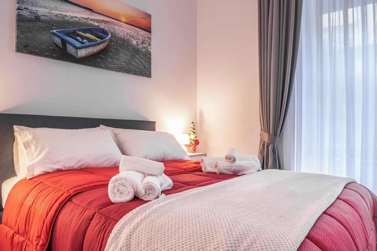 A neatly made bed with red bedding, white pillows, and rolled towels. A painting of a boat on water is on the wall above the bed.