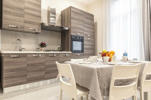 A modern kitchen with wooden cabinets, a stainless steel range hood, and a built-in oven. There is a dining table with chairs and a fruit bowl on the table. - Casina Rossa (Naples)
