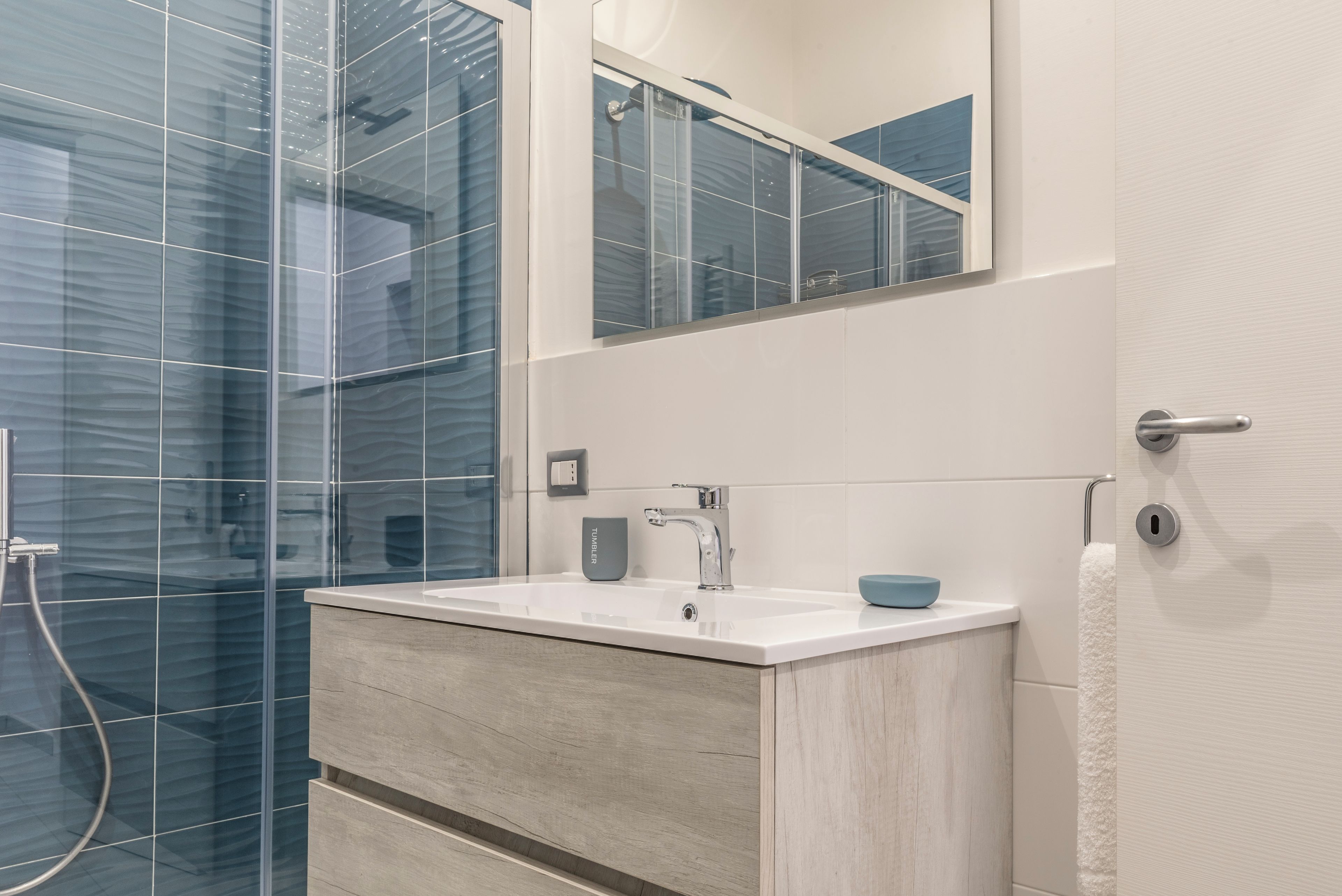Une salle de bain moderne avec une douche à parois de verre, un lavabo blanc avec un porte-savon bleu et un meuble-lavabo en bois.
