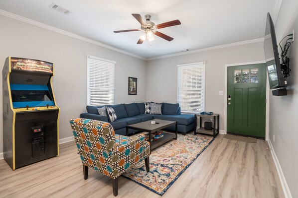 A living room with a blue sofa, patterned armchair, coffee table, arcade machine, and a ceiling fan. - NEW! UPTOWN Cottages - 2bed/1bath - new furnishings! (Hot Springs)