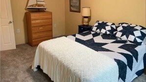 Une chambre à coucher avec un lit, une commode, une lampe et une maquette de voilier posée sur la commode.