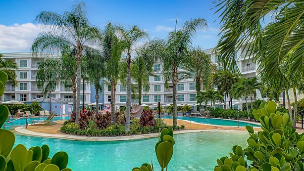 Ein Resort mit einem zentralen Pool, umgeben von Palmen und einem mehrstöckigen Gebäude mit Balkonen.