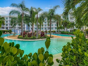Un resort con una piscina central rodeada de palmeras y un edificio de varios pisos con balcones.