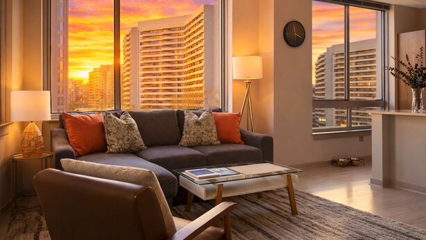 A modern living room with a sofa, a coffee table, and a chair. There are two lamps, a clock on the wall, and a view of tall buildings through large windows. - Wow! 5 min to DC/Metro/Mall Stunning 2B King Bed Pool Gym (Arlington)
