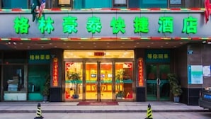 A hotel entrance with a green signboard, glass doors, and a red mat at the entrance. There are potted plants on either side of the entrance. - Greentree Inn Zhonggang City Express Hotel (Jiaxing)
