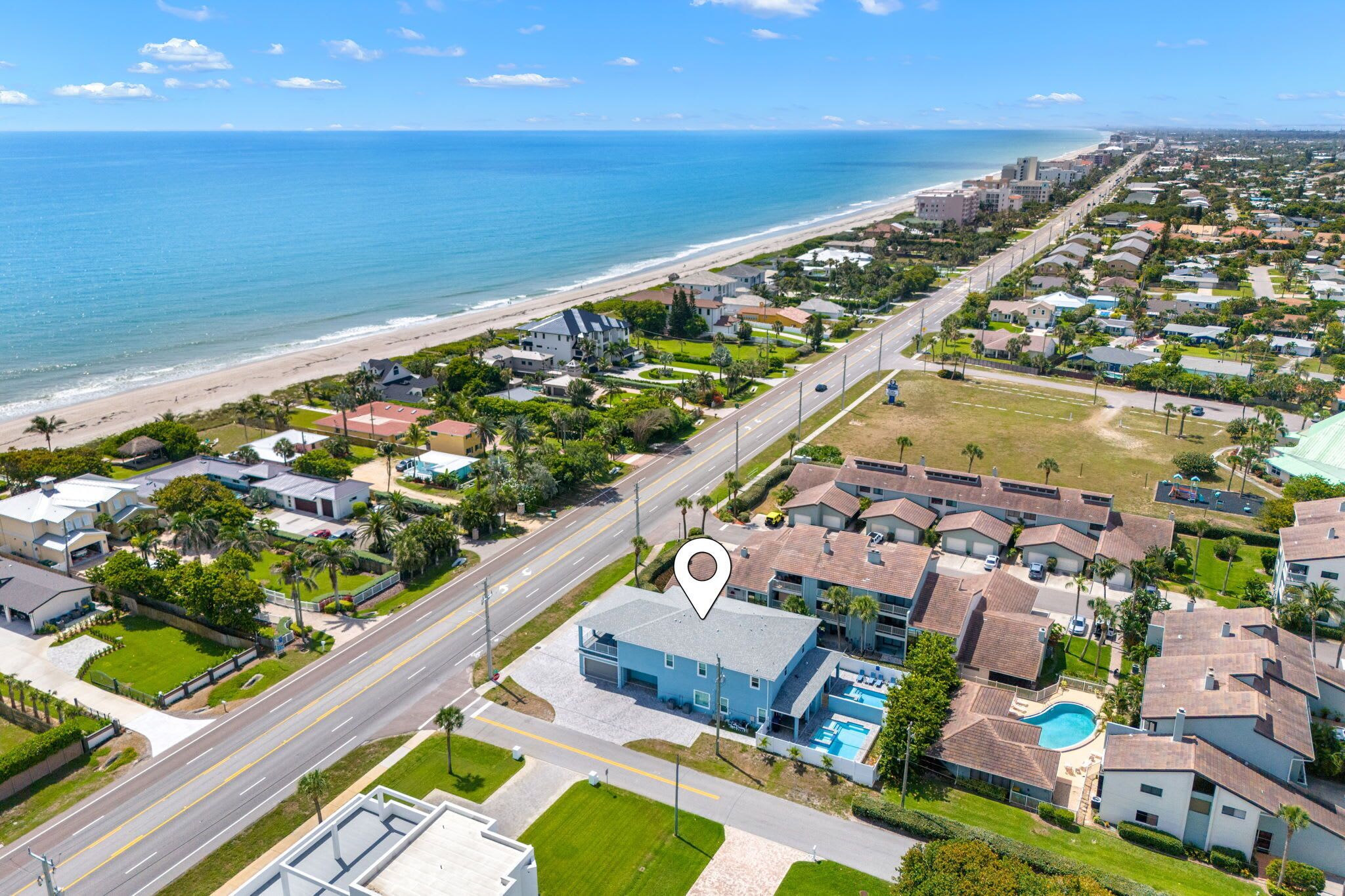 Uma área residencial costeira com uma grande casa azul com piscina, rodeada por outras casas e uma praia ao fundo.