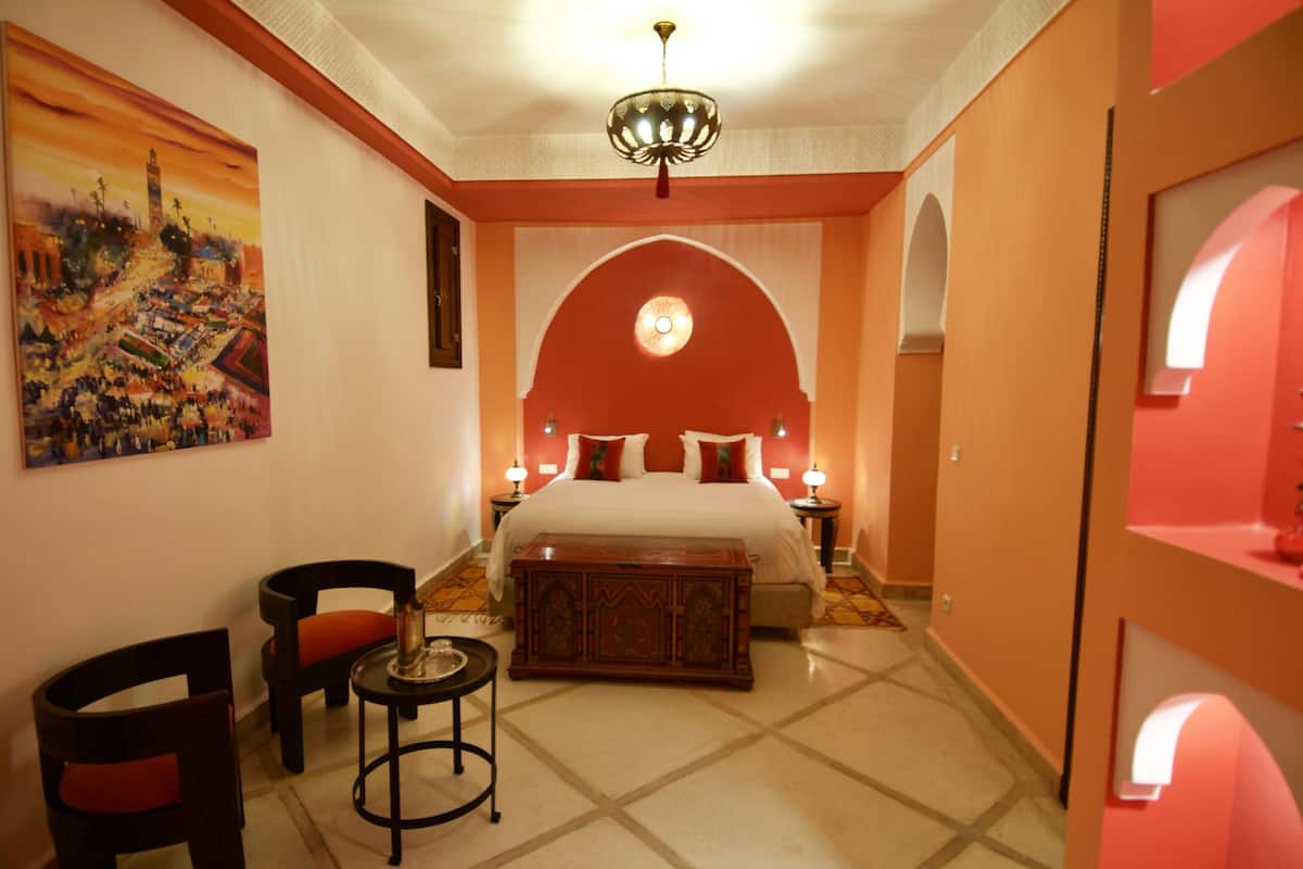 A hotel room with a bed, a nightstand, a chair, a small table, and a painting on the wall.