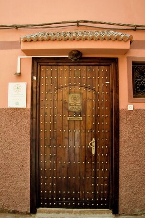 A wooden door with a metal latch and a decorative plaque, set in a building with a textured pink wall and a tiled roof. - La Claire Fontaine (Marrakech)