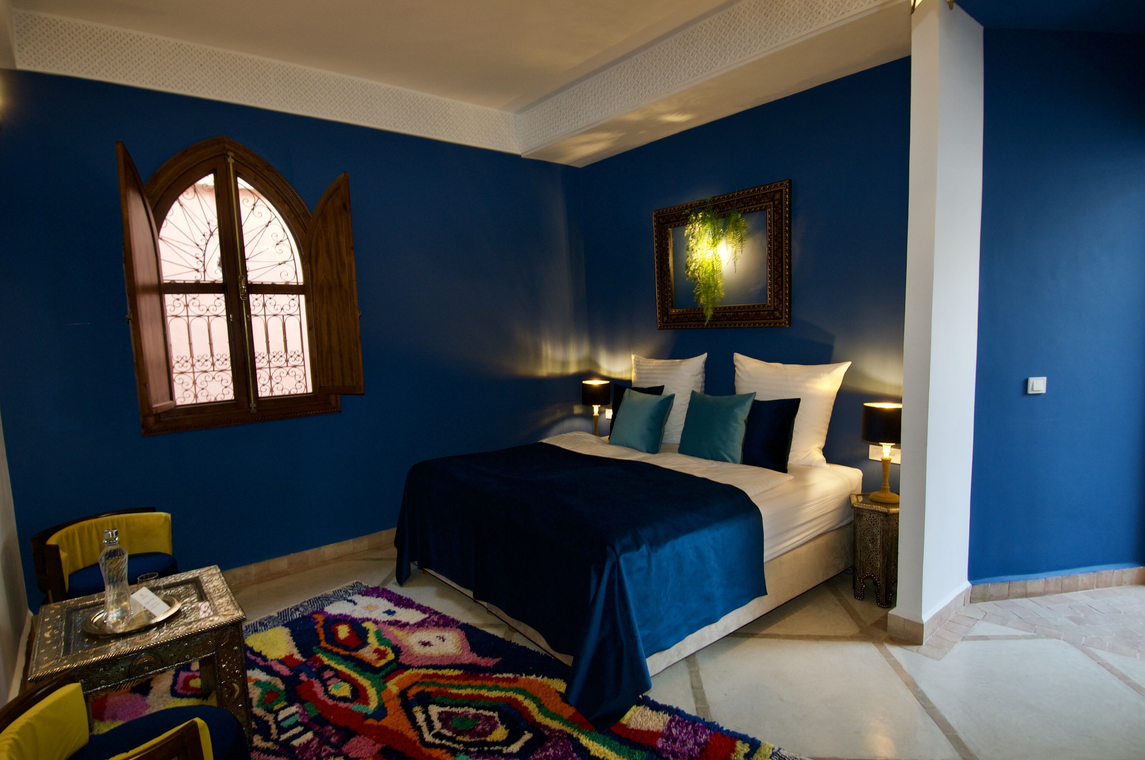 A bedroom with a large bed, a window with decorative iron bars, a small table with a vase, and a colorful rug.