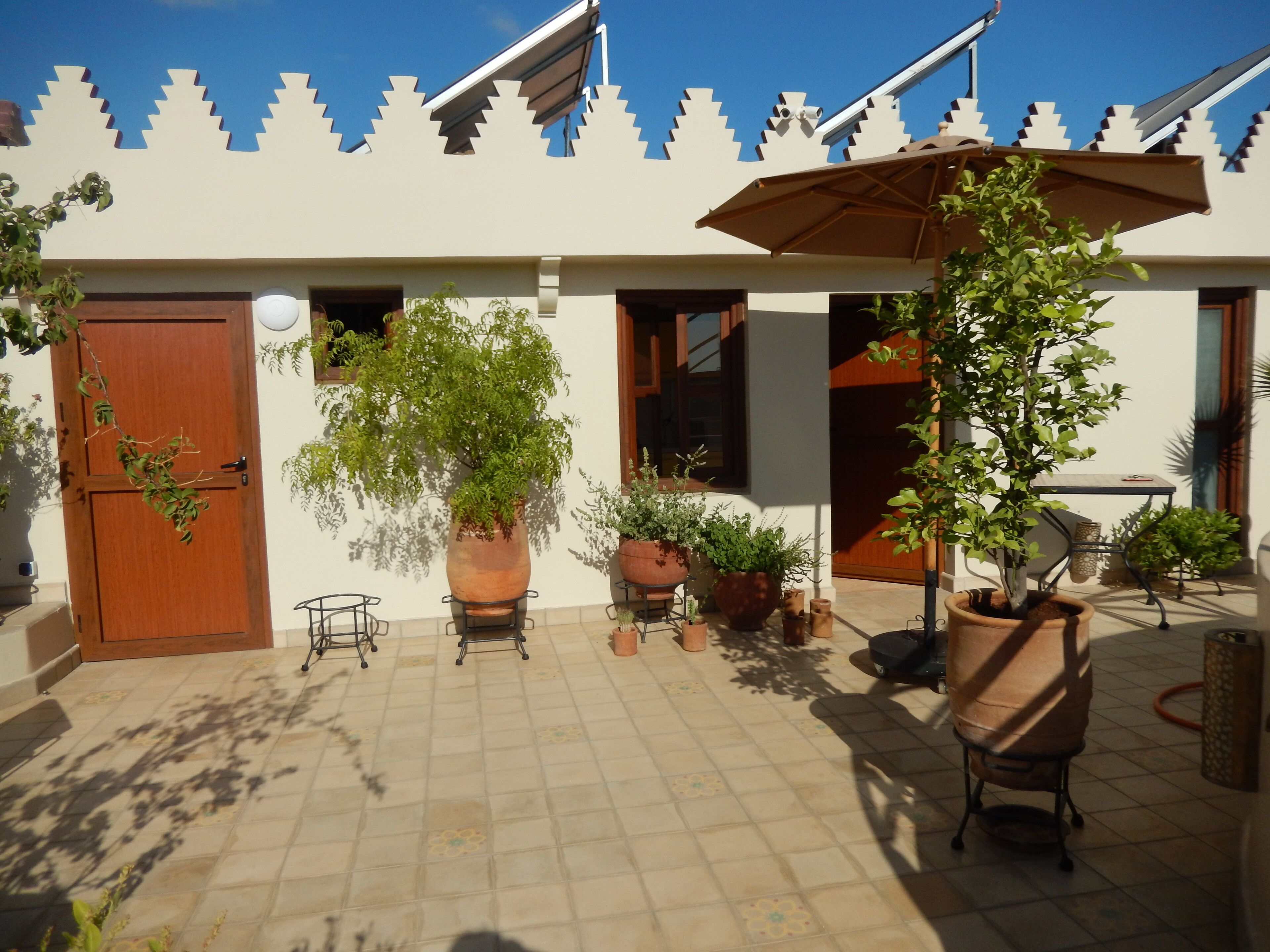 A courtyard with a tiled floor, potted plants, a wooden door, and a patio chair under an umbrella.