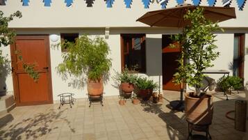 A courtyard with a tiled floor, potted plants, a wooden door, and a patio chair under an umbrella.