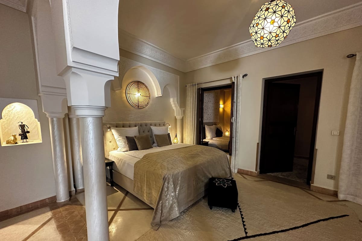 A spacious bedroom with a large bed, a decorative wall light, and a view into an adjacent room.