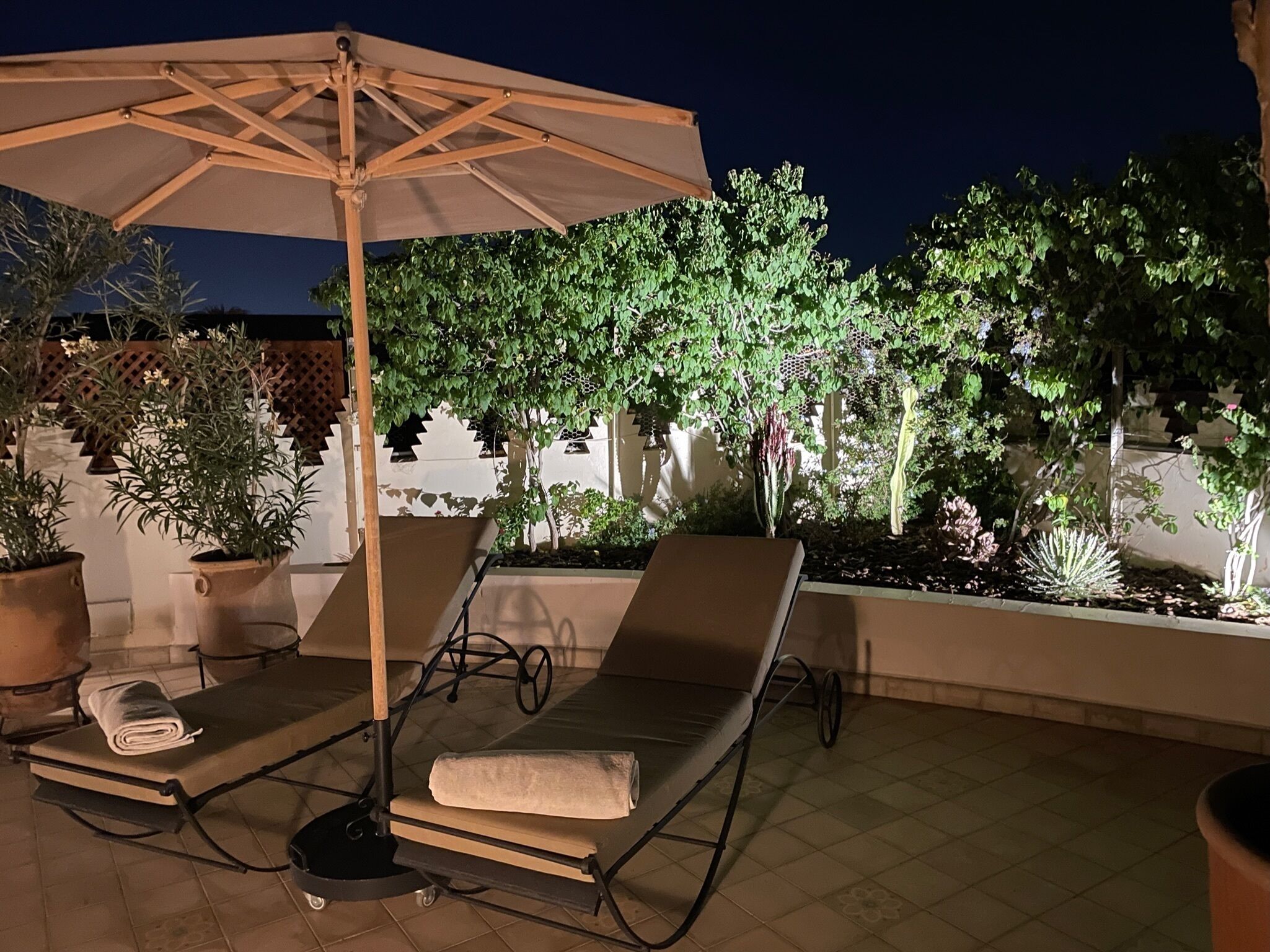 A rooftop terrace with a lounge chair and an umbrella, surrounded by potted plants and a white wall.