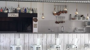 Zona de bar con taburetes blancos, variedad de botellas y vasos en estanterías, luces colgantes y un letrero que dice "Boliche de Amigos".