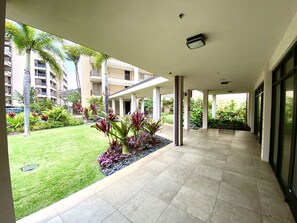 A covered walkway with a tiled floor, surrounded by greenery and palm trees, leading to a building with multiple balconies.
