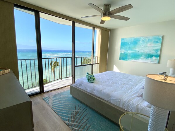 A hotel room with a bed, a ceiling fan, a large window with a view of the ocean, and a painting on the wall.