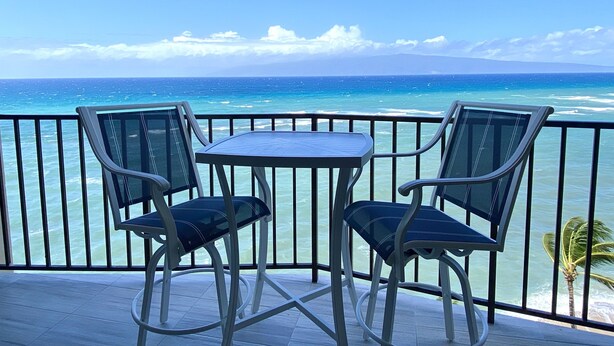 Een balkon met twee stoelen en een tafel, met uitzicht op een turquoise oceaan met golven en een heldere lucht.